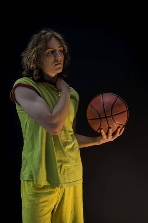 Young boy with long blond hair wearing a green jersey caressing his shoulder and holding a basketball.の写真素材