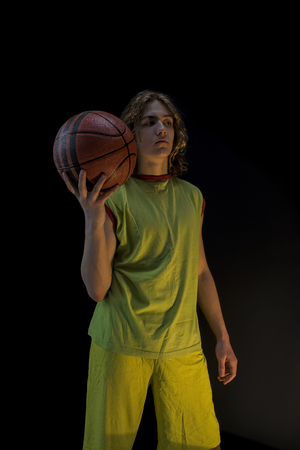 Young boy with long blond hair wearing a green jersey standing and holding up a basket ballの写真素材