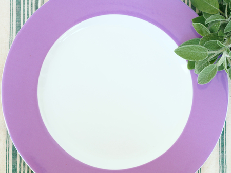 Home garden sage herb sprout near an empty white plate with purple edge on green and white table cloth, top viewの写真素材