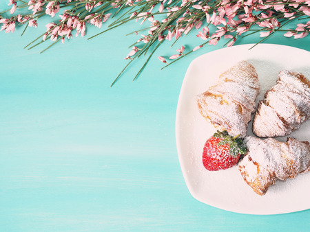 Spring green pastel wooden background with pink flowers at the top and croissants with a strawberry on the right. toned imageの写真素材