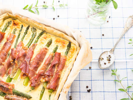 Savory pie with asparagus, prosciutto, marjoram and egg and cream mixture in a baking tin with parchment on checkered table clothの写真素材