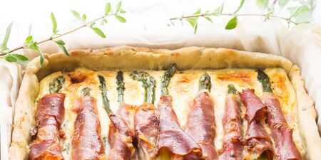 Savory pie with asparagus, prosciutto, marjoram and egg and cream mixture in a baking tin with parchment on checkered table cloth, closeup bannerの写真素材
