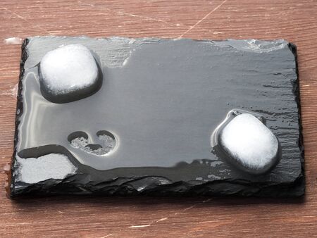 Wet black slate plate with ice cubes and a heart shaped spot on the surface. Dark wooden background. Copy spaceの写真素材