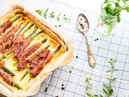 Quiche with asparagus, prosciutto and marjoram herb in a square baking tin on checkered table clothの写真素材