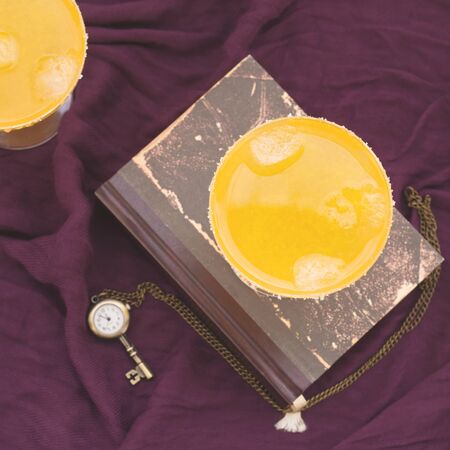 Two orange alcohol cocktails on purple textile with a book. Top view