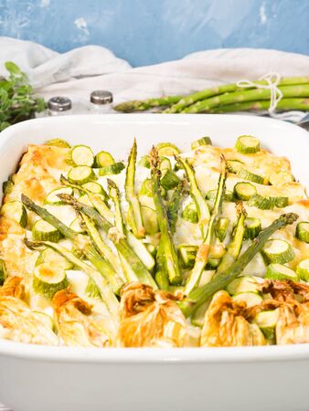 Home made asparagus lasagna casserole with zucchini, ricotta, mozzarella cheese and marjoram herb in a ceramic tin on wooden table. Rustic spring baked one plate food