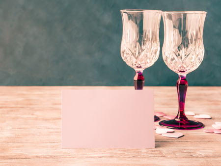 Two stemmed champagne glasses with pink hearts blank paper note card to fill with your text on wooden textured background. Valentines day wedding romantic date invitation. Tonedの写真素材