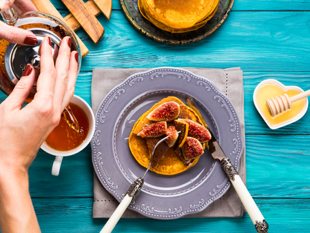 Eating pumpkin pancakes with figs and honey with hot tea. Autumn food background on green wooden table. Woman's handsの写真素材