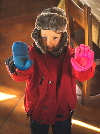 Little 5-year-old boy in snowsuit and cap with earflaps making funny face expressions with different mittensの写真素材