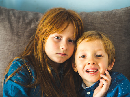 Two blonde sibling kids in blue shirts on a sofa hugging. Portrait of children indoorsの写真素材