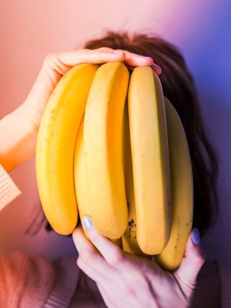 Girl covering her face with bunch of bananas. Multi color neon background with soft light. Pink and violetの写真素材