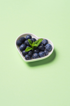 Fresh blueberries in heart shaped plate on pastel green background. Healthy breakfast conceptの写真素材