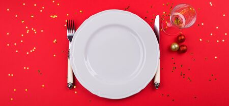 Christmas New year dinner concept color flat lay with empty white dish and silverware, champagne flute on red monochrome background with golden confetti. Festive bannerの写真素材