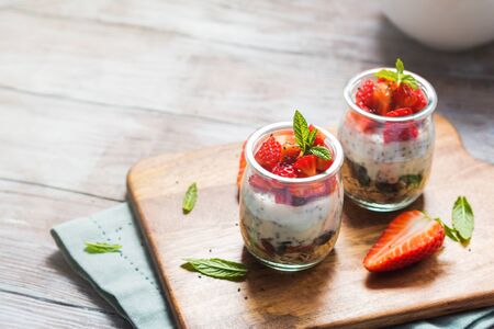 Yogurt granola parfait with strawberries and chia seeds. Healthy single portion breakfast served in glass cups on wooden board with green napkin. Natural wooden table backgroundの写真素材