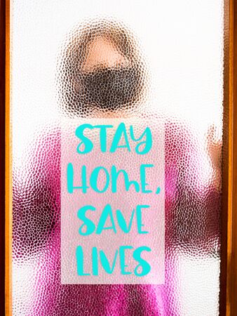 Woman wearing black face mask behind door glass at home during Coronavirus pandemic quarantine. Stay home, save lives textの写真素材