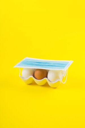 White and one brown egg under medical face mask on yellow bold color paper background. Easter, art...