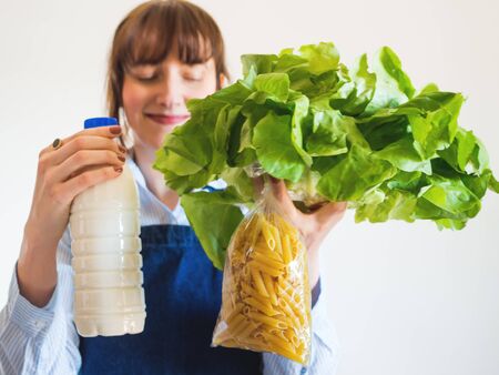 Delivery girl or small shop owner wearing blue apron holding food staples - fresh vegetables, pasta and milk - in gloved hands. Food delivery, grocery shoppingの写真素材