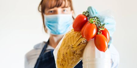 Delivery girl or small shop owner wearing protective face mask holding food staples - fresh vegetables, pasta and milk - in gloved hands. Safe delivery, grocery shopping during coronavirus pandemicの写真素材