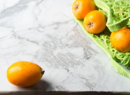 Loquat Japanese medlar fruit in green mesh shopping bag on marble table. Zero waste produce shoppingの写真素材