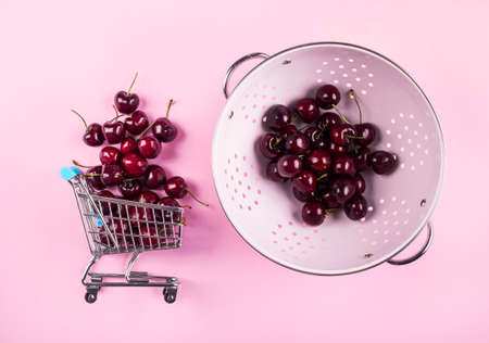 Cherries in toy shopping cart and pink kitchen colander on pink background. Fresh summer produce dlivered to your home concept, flat layの写真素材