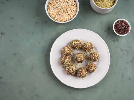Home made vegan energy protein balls with oats, nuts, dates, dried fruit, flax and hemp seeds, chocolate nibs and maple syrup on white dish over green textured background. Flat layの写真素材