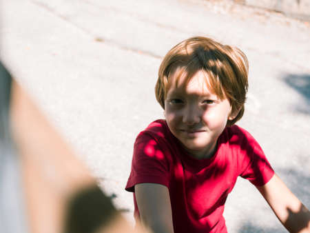Little blonde boy wearing red T-shirt riding a bike outdoors in summer on a sunny day. Authentic...