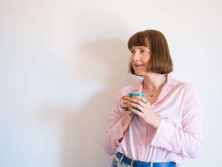 Young woman wearing jeans and pink shirt drinking coffee and smiling over white wall background.の写真素材