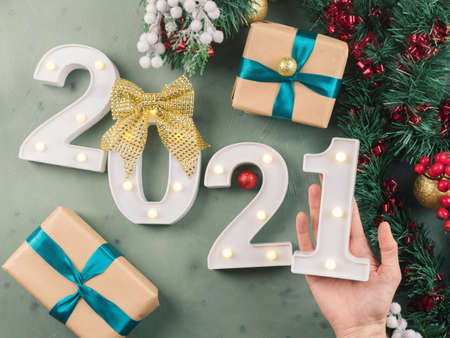 New year festive green background with 2021 illuminated numbers with ornament and wrapped gift boxes on dark green. Female hand holding number 1.の写真素材