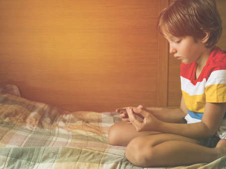 Little blonde boy playing games at smartphone sitting on his bed.の写真素材