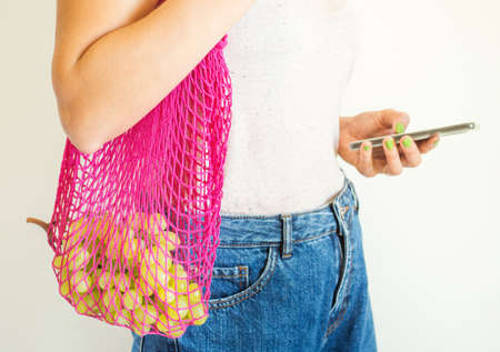 Young woman with pink reusable mesh bag with fresh produce and smartphone in hand. Zero waste,...