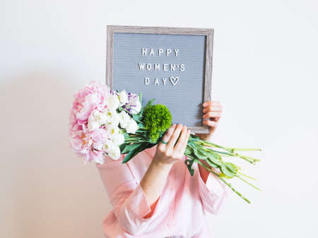 Young woman holding pink bouquet and letter board saying happy womens day on gray wall background