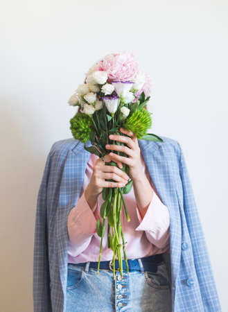 Young woman wearing blue jacket and pink shirt holding beautiful pastel peony bouquet in front of face on gray backgroundの写真素材