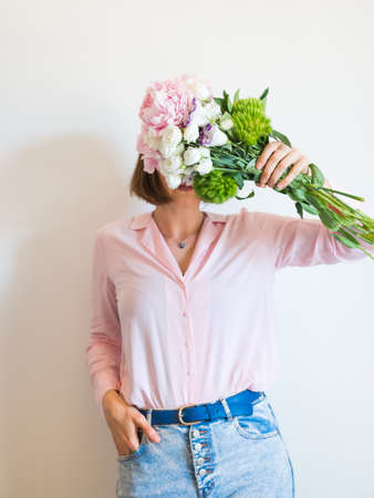 Young woman holding beautiful pink bouquet in front of her face on light gray background. Faceless portraitの写真素材