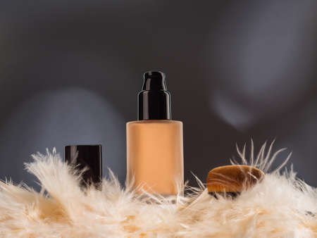 Unbranded generic foundation bottle with feathers and professional brush on dark gray background with shadows. Advertising display on podiumの写真素材