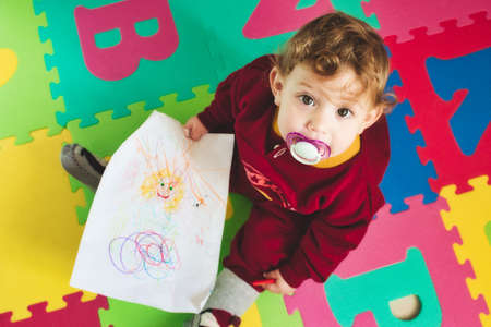 Cute baby girl with dummy on alphabet play mat drawing on paper with a colored crayon