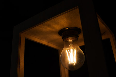 Close-up of warm yellow glowing light bulb encased in a wooden box against a black backgroundの写真素材