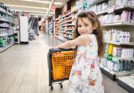 A cute young girl pushing a small shopping trolley in a supermarket aisle looking back at the camera and smilingの写真素材
