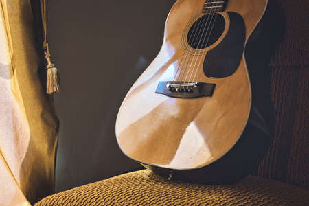 A Spanish acoustic classical guitar with steel and nylon strings resting on a chair next to a window...