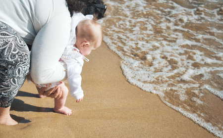 Barefoot mother and baby daughter on the sea shore on a sandy beach walking close to the surf foam on the wavesの写真素材