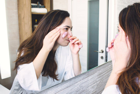 An attractive white Caucasian woman using a jade roller on her face in front of the bathroom mirrorの写真素材