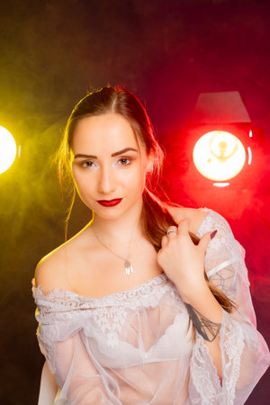 A stunning young European woman strikes a pose in a studio, bathed in a dynamic blend of red and yellow light emanating from behind. The warm hues accentuate her beauty and lend an artistic flair to the image.の写真素材