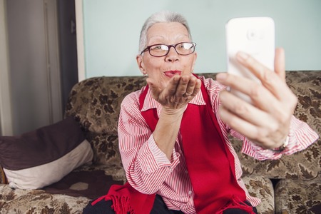 Senior old woman sending love and kisses to her family over social media apps using her cellphoneの写真素材
