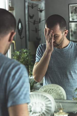 Man standing in front of a mirror, sad and depressed, experiencing a negative self imageの写真素材