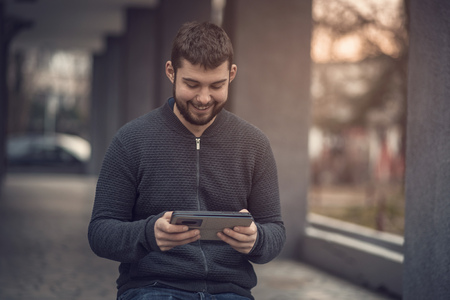 Young mid-twenties man sending a text message on his tablet, speaking with a friend as hes taking a...