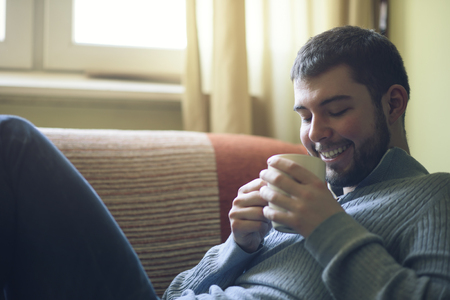Young bearded man laying on his sofa, enjoying the smell of his morning coffee or teaの写真素材