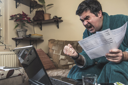 Enraged mature man yelling at his bills and financial reports, dressed in bathrobe, sitting in his living roomの写真素材