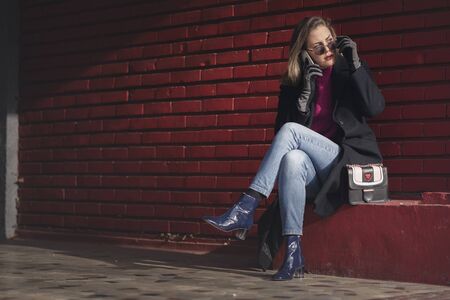 Unique stylish and classy young lady using her phone outdoors, sitting next to a red brick wallの写真素材