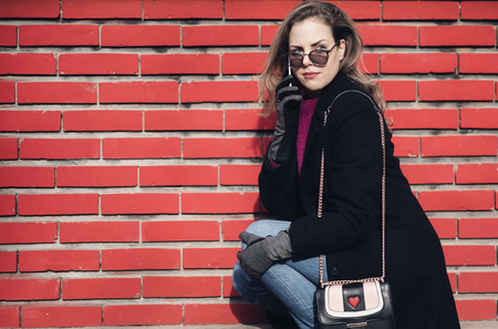 Unique stylish and classy young lady using her phone outdoors, sitting next to a red brick wallの写真素材