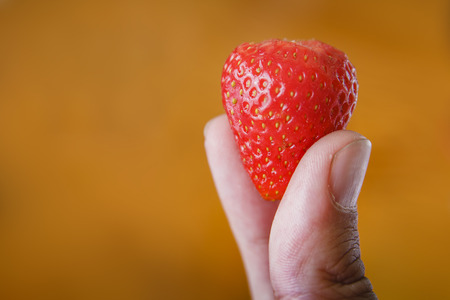Two fingers holding a strawberry against wooden yellow background, no petals, pealedの写真素材