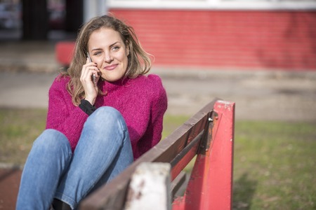 Sweet young woman sitting on the bench in a park, having a wonderful conversation on her phone, feeling free and relaxedの写真素材